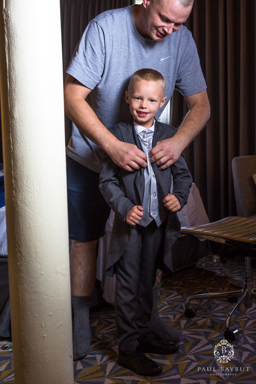 A man in casual clothes helps straighten a young boy's formal suit and tie.
