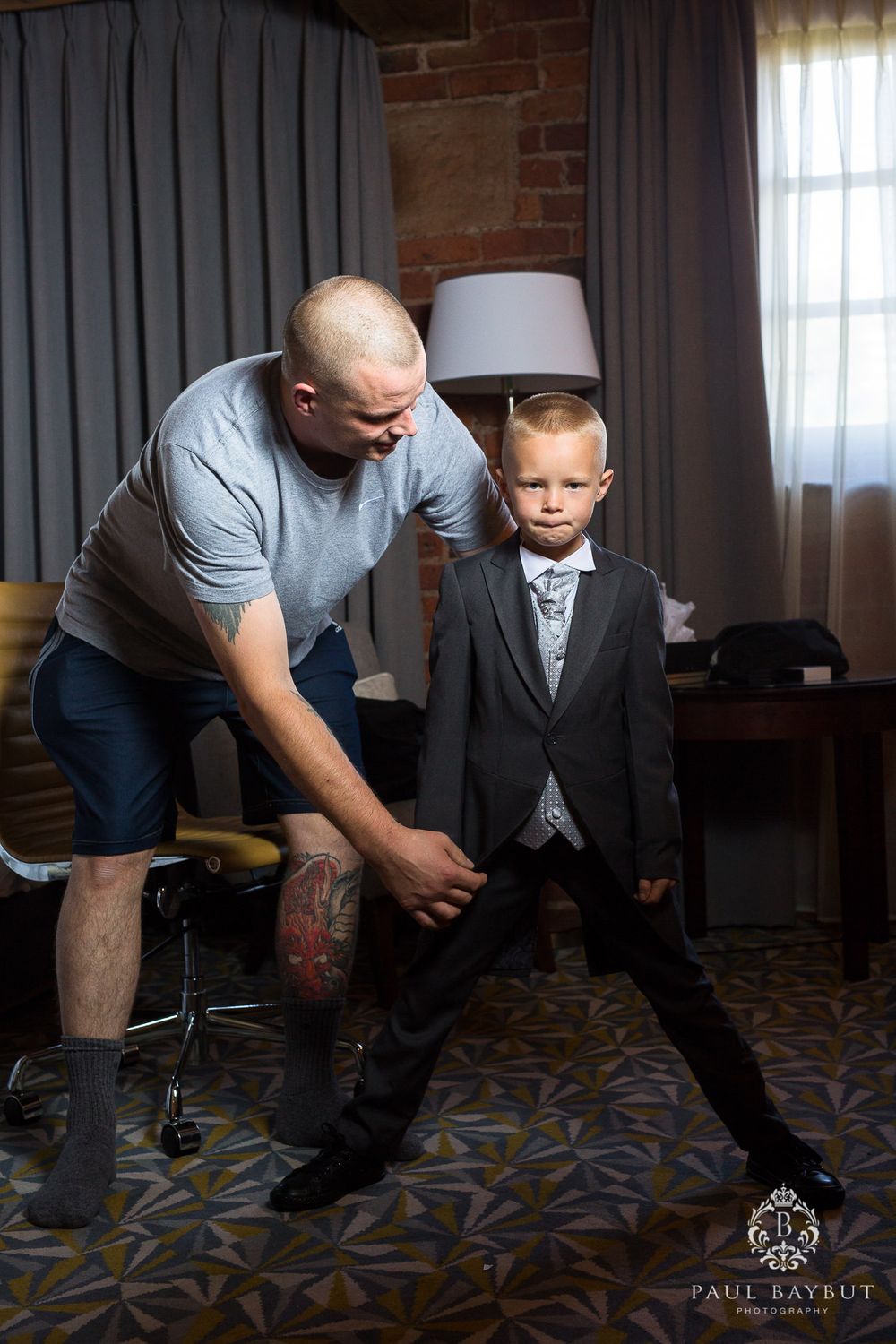 A man assists a young boy with his formal suit while standing in a room with brick walls.
