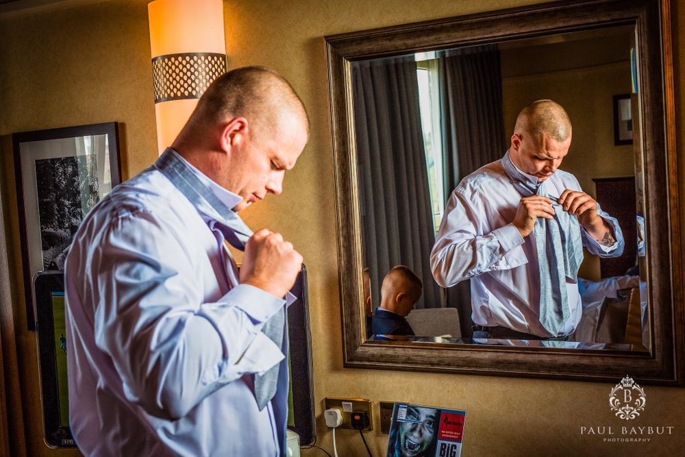A man in a light blue dress shirt adjusts his tie while looking in a mirror.
