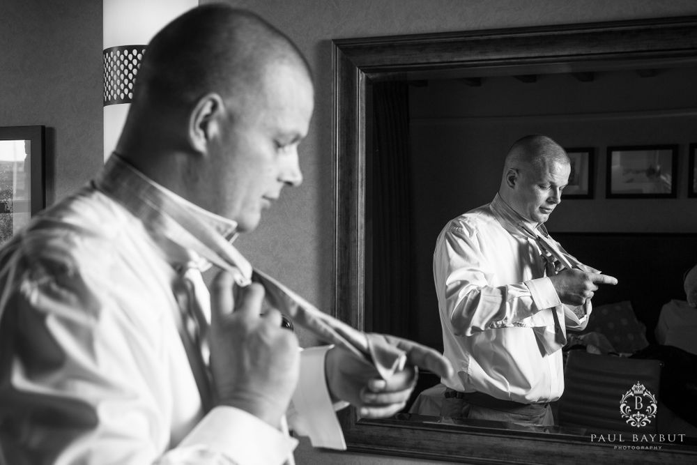 A black and white image of a man adjusting his tie while looking in a mirror.
