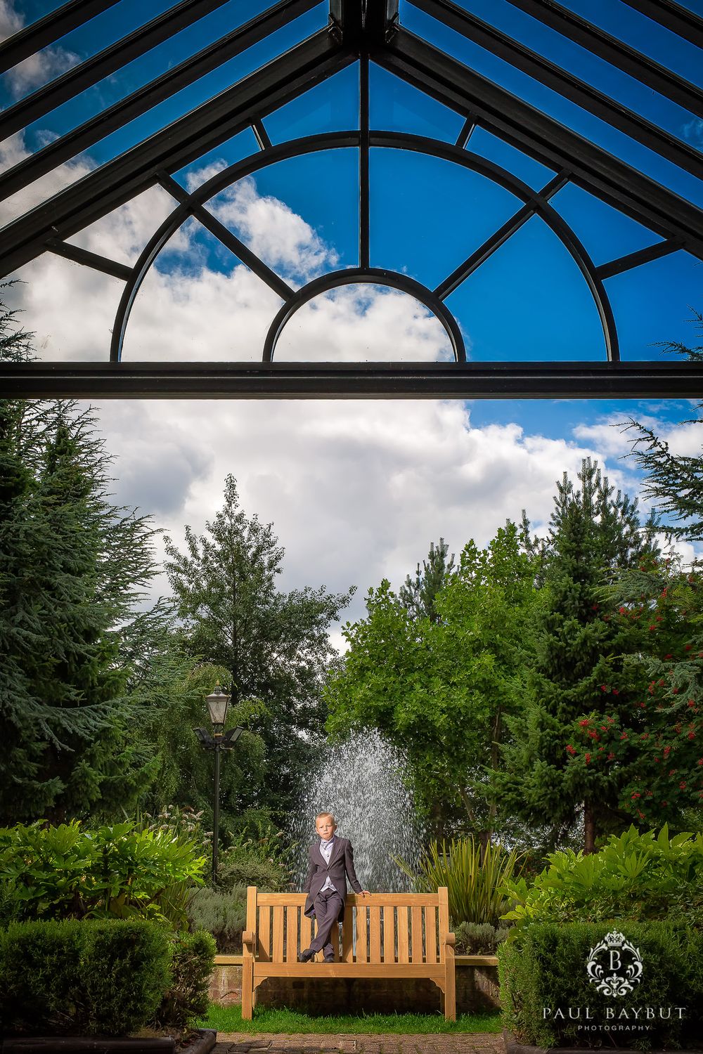 Garden bench under a glass conservatory roof with fountain and greenery visible through the windows.