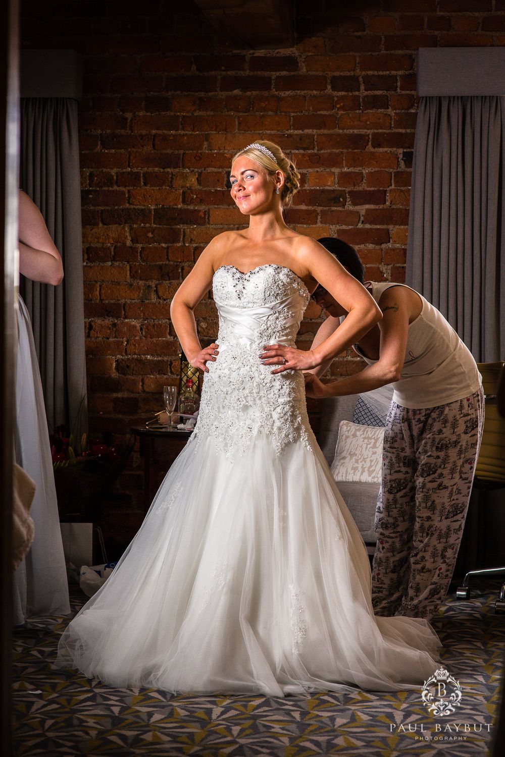 Wedding dress fitting in a rustic brick-walled room with dramatic lighting.