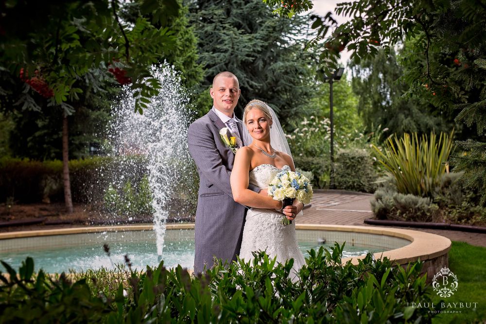 Couple poses by garden fountain in wedding attire with lush greenery background.