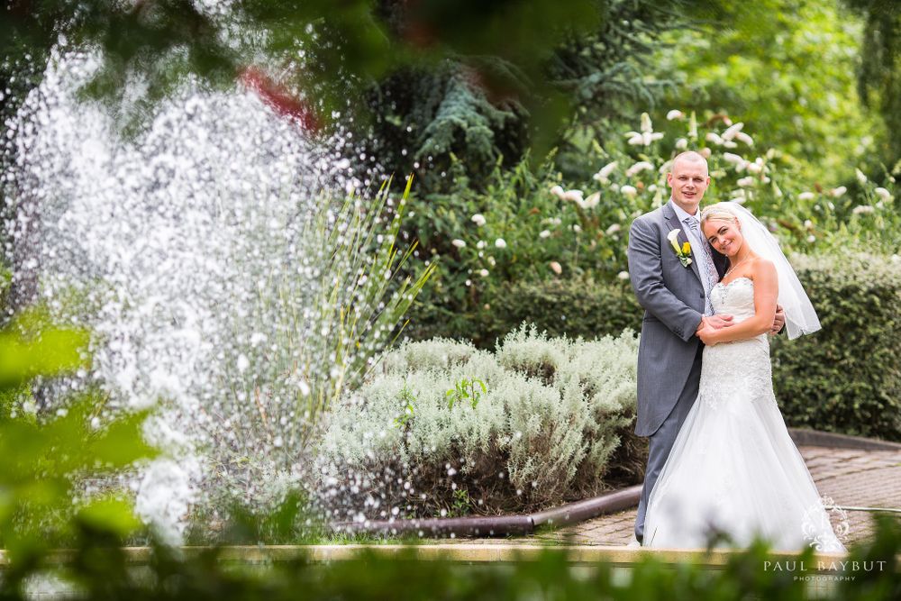 Wedding couple stands near fountain in beautiful garden setting with water spray.