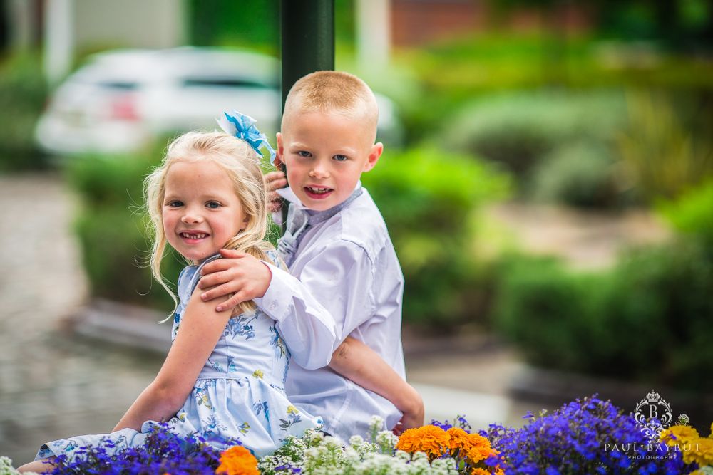 Two children pose together among colorful purple and orange flowers in a garden setting.