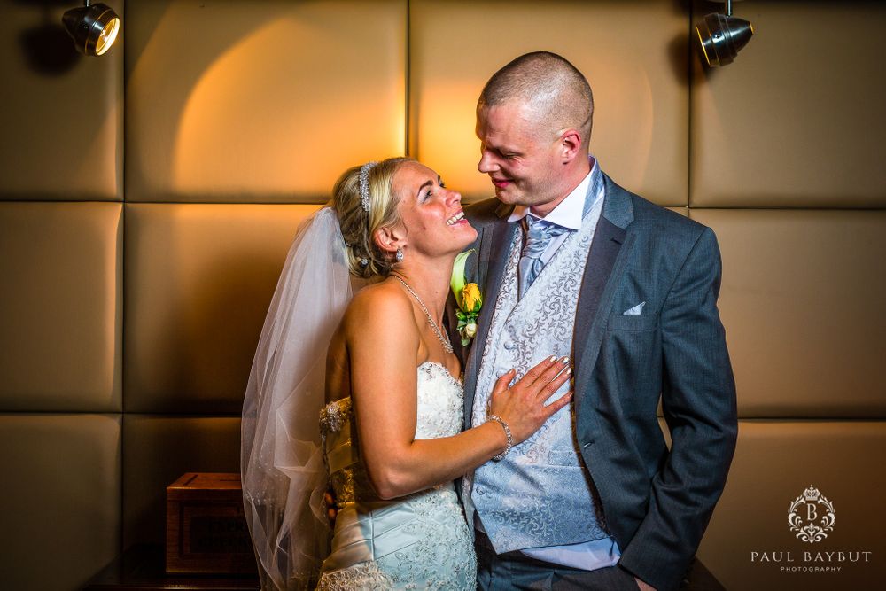 A romantic wedding portrait shows a couple sharing an intimate moment against a warm-lit padded wall.
