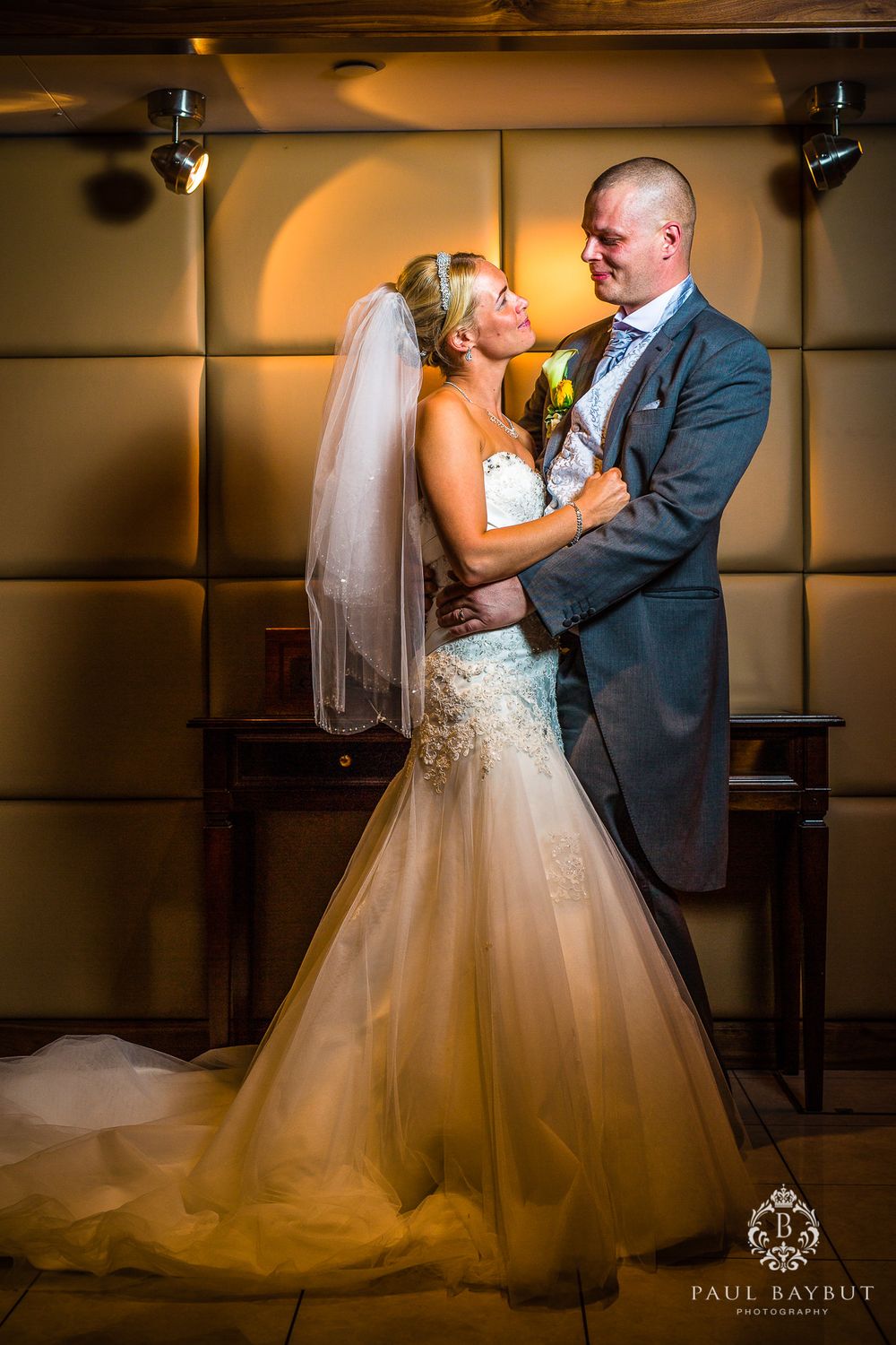 A full-length portrait of newlyweds in elegant attire sharing a romantic moment against a warm-lit backdrop.
