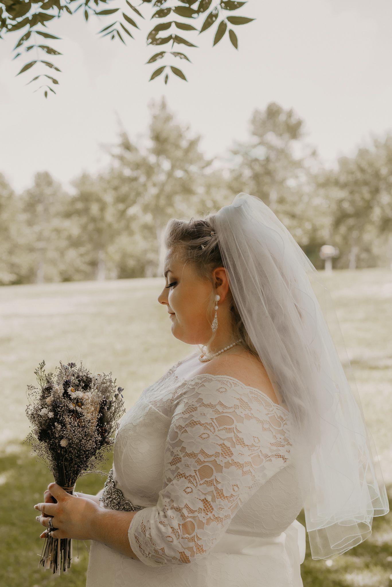 Bride in lace dress holds dried flower bouquet in outdoor garden setting.
