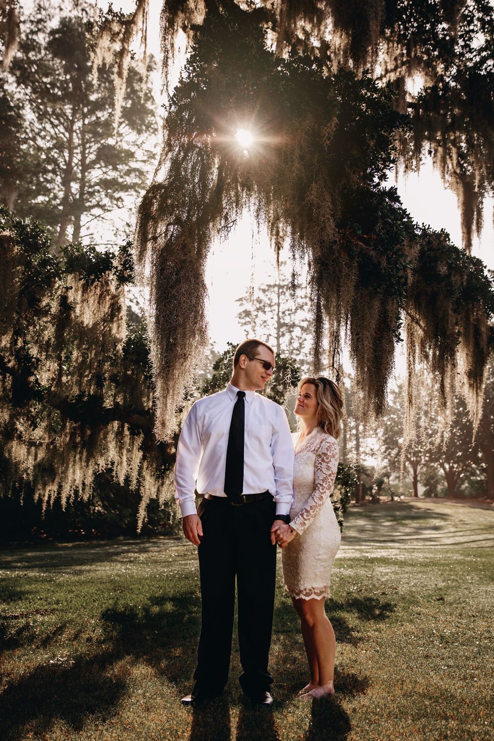 Couple stands together beneath Spanish moss hanging from trees during golden hour.