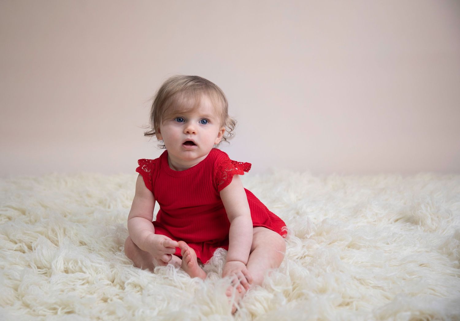 girl sitter session, sitting on cream flokati, cream vinyl backdrop, child wearing red bodysuit.