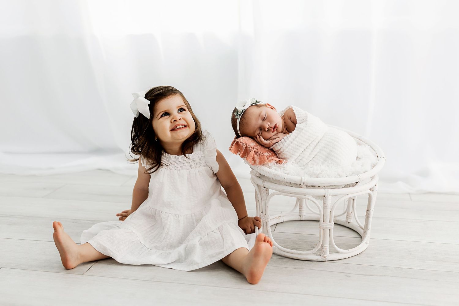 Children in white dresses pose with wicker chair in bright studio setting.