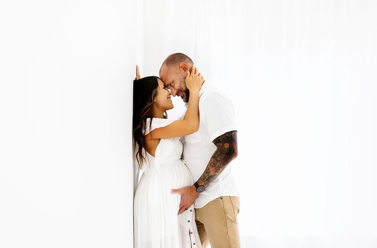 Couple in white outfits sharing intimate moment against white wall.