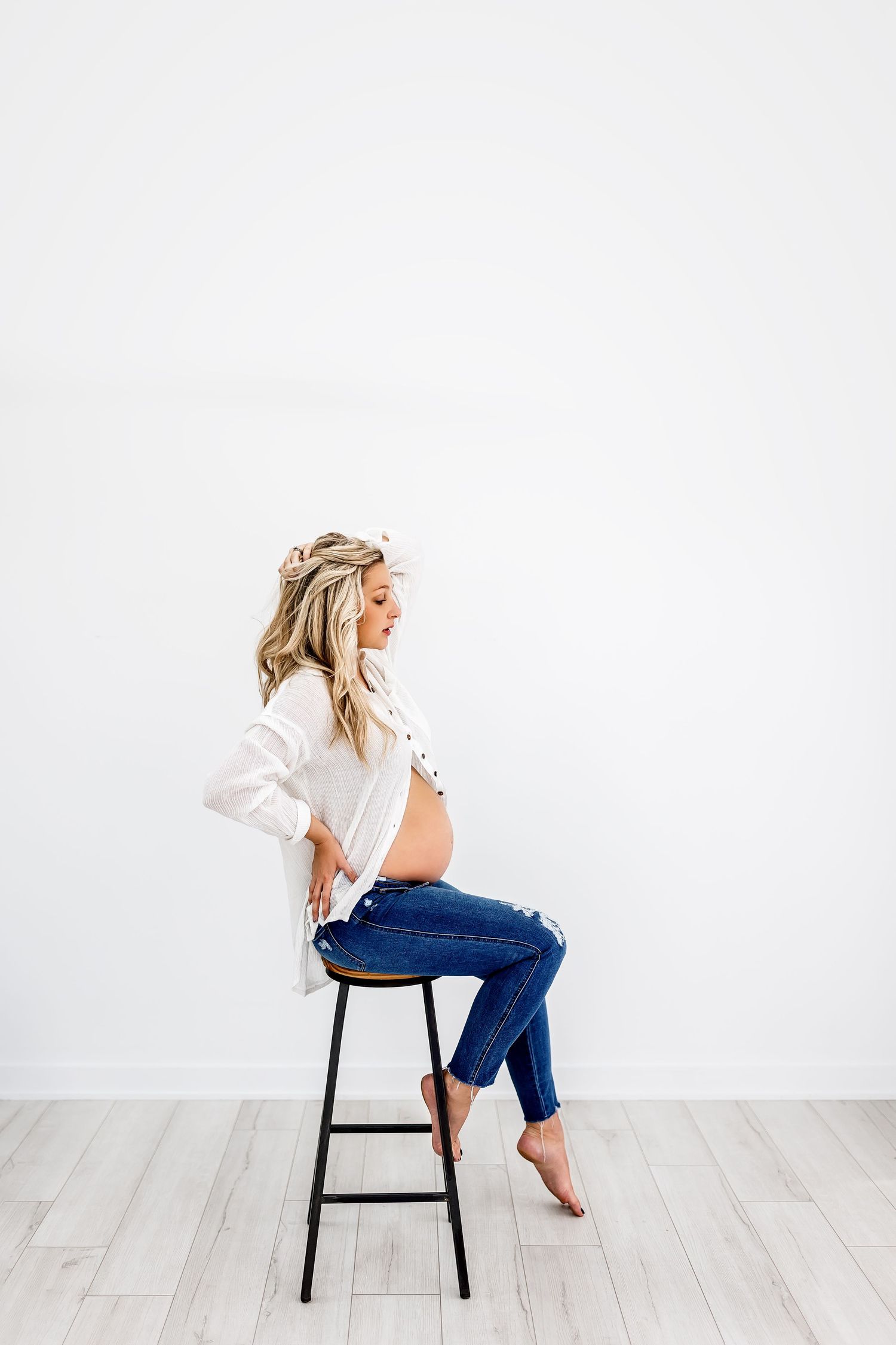 Elegant maternity portrait of woman in white top and jeans sitting on black stool.