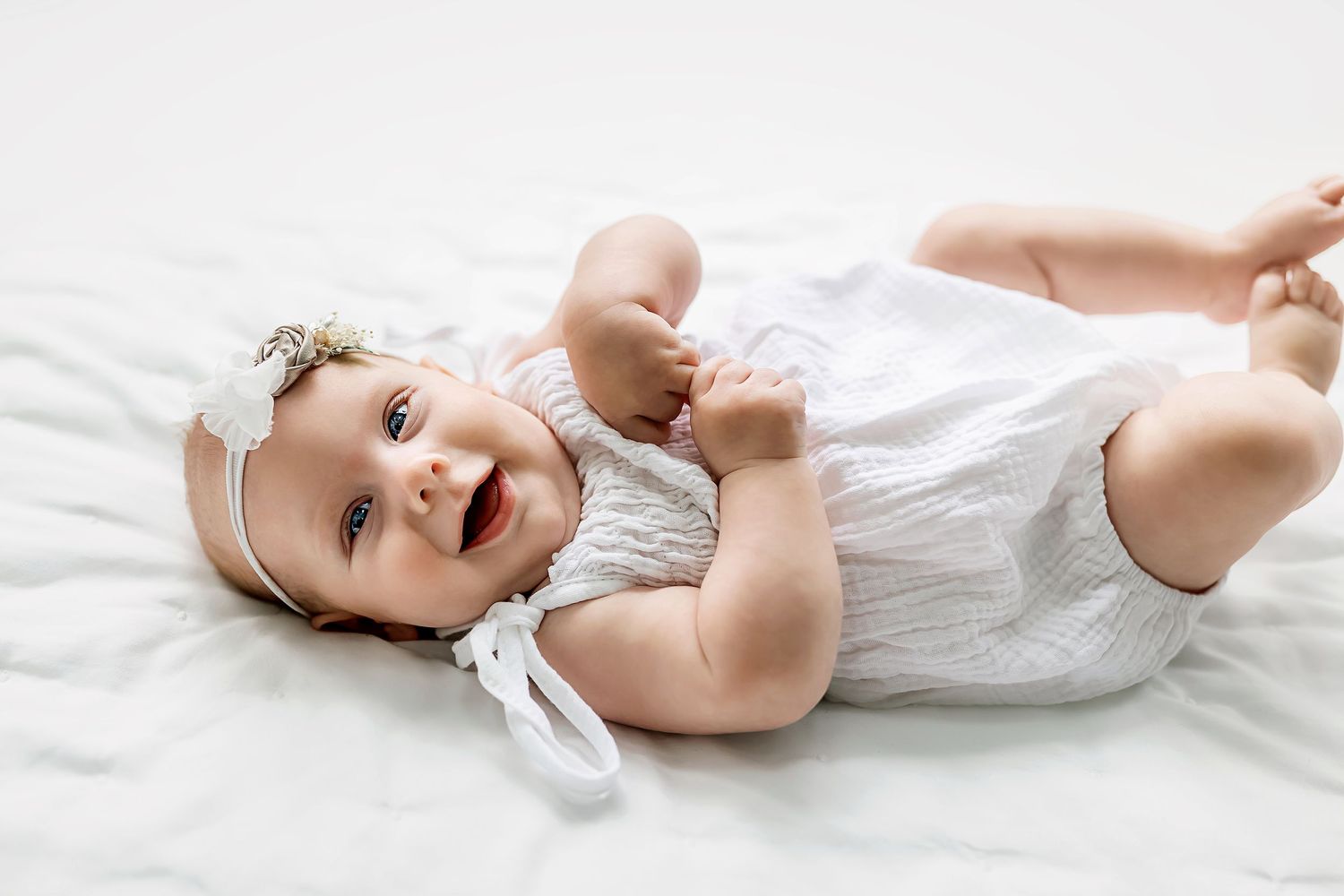 Adorable baby in white dress lying on bed with lace headband.
