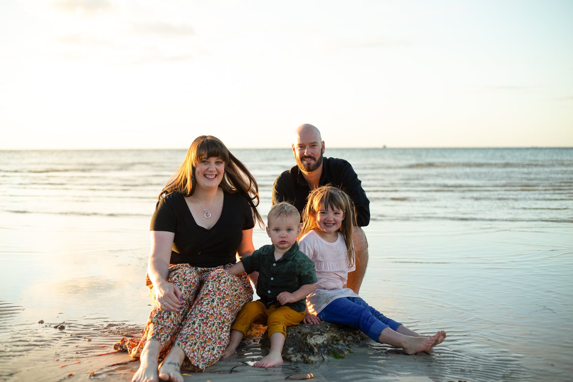 Bianca Brook, husband and children at the beach sitting on a rock with the sunset behind them smiling