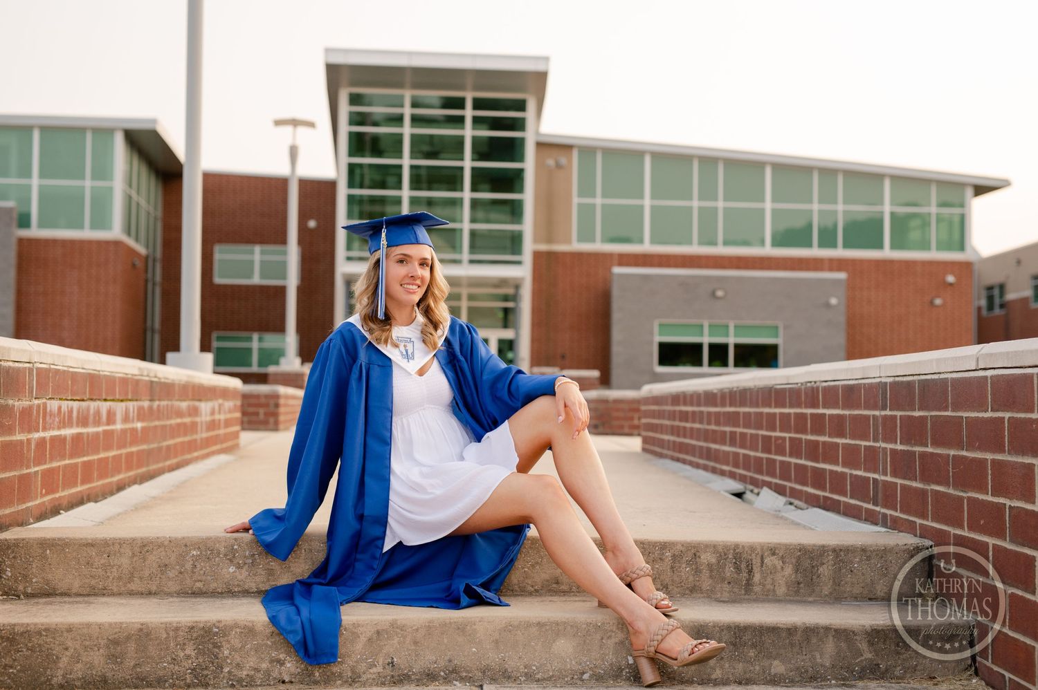 Graduate in blue cap and gown poses on brick steps near a glass-fronted building.