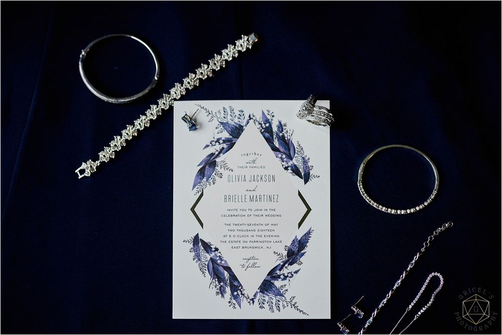 Wedding invitation with blue feather design surrounded by jewelry and accessories on dark fabric.