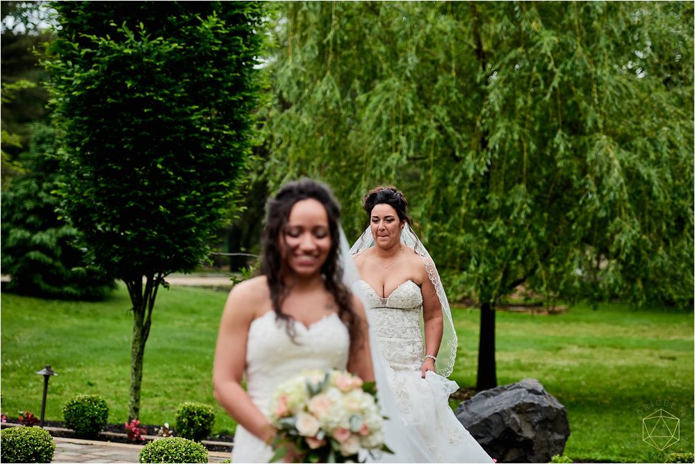 Two brides in white wedding dresses posing in a garden setting with willow trees.
