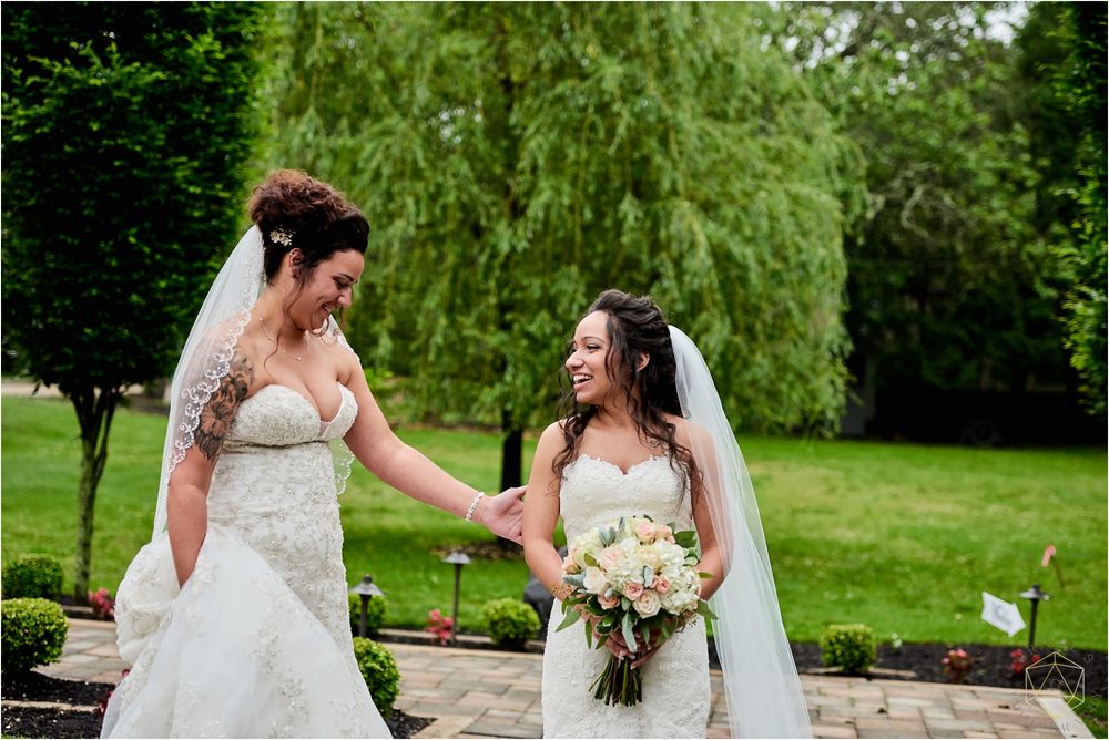 Two brides in white gowns sharing a happy moment in a garden setting.