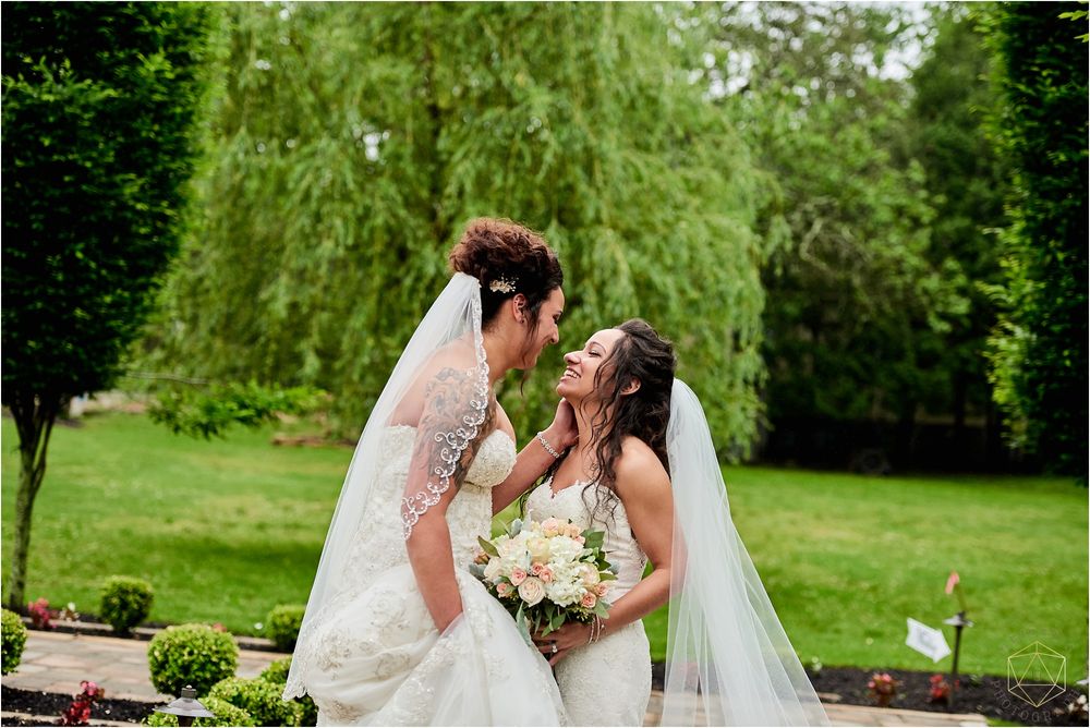 Two brides share a joyful moment together under a weeping willow tree in a garden setting.