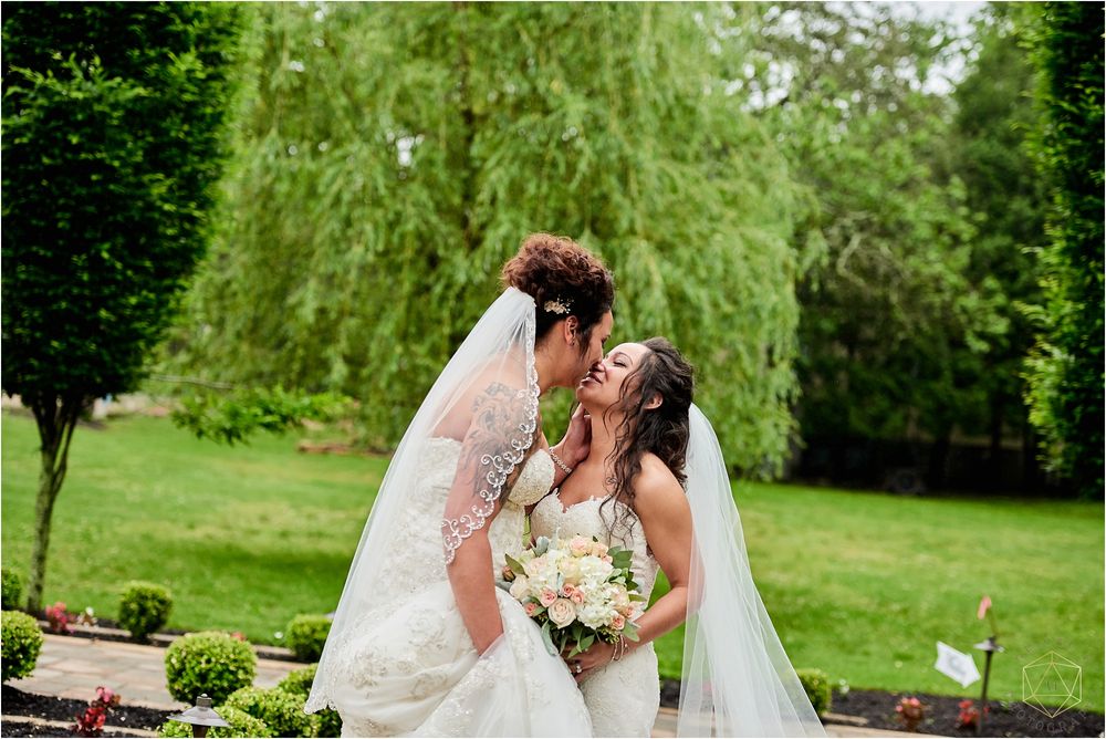 Two brides laugh together while holding a white floral bouquet in an outdoor garden.