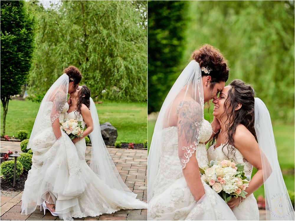 Two brides in white gowns embrace on a stone patio with weeping willows in the background.
