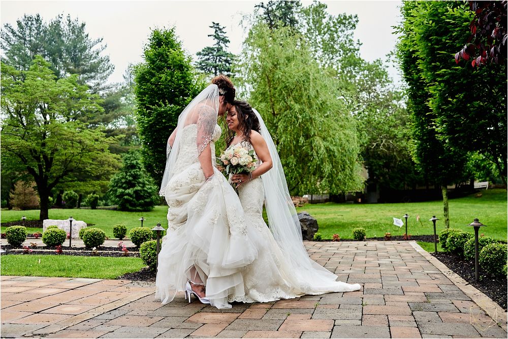 Two brides in flowing white dresses pose together on a stone patio surrounded by lush gardens.