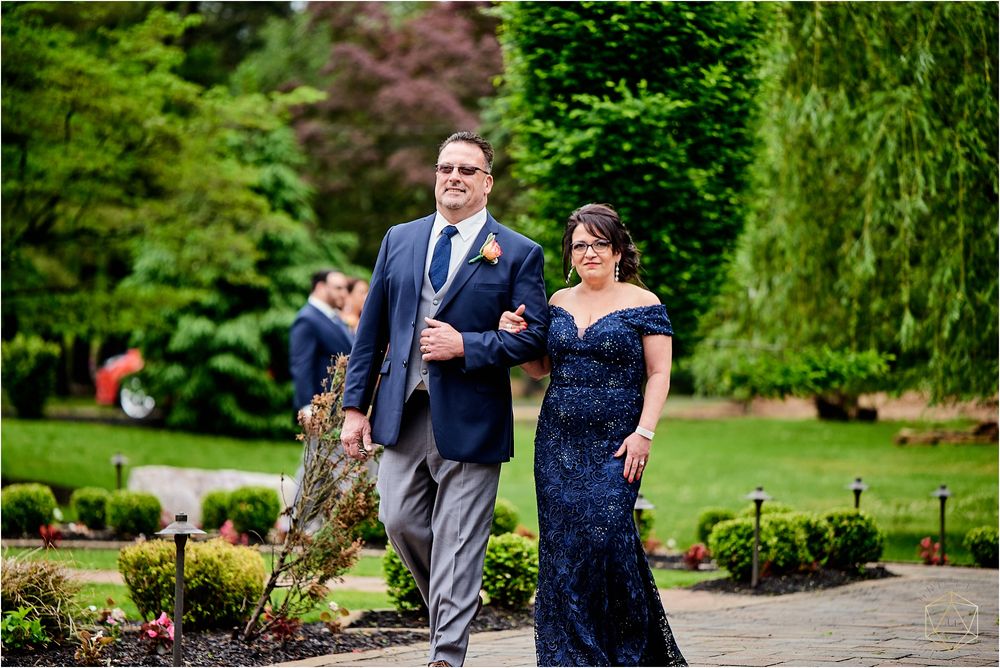 Wedding party members walk down a garden path in formal navy and grey attire.