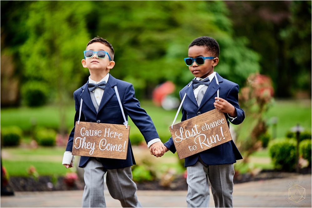 Two young ring bearers in suits and sunglasses walk together holding wooden signs.