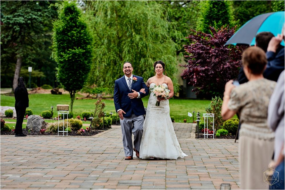 A wedding processional takes place on a stone patio surrounded by garden landscaping.
