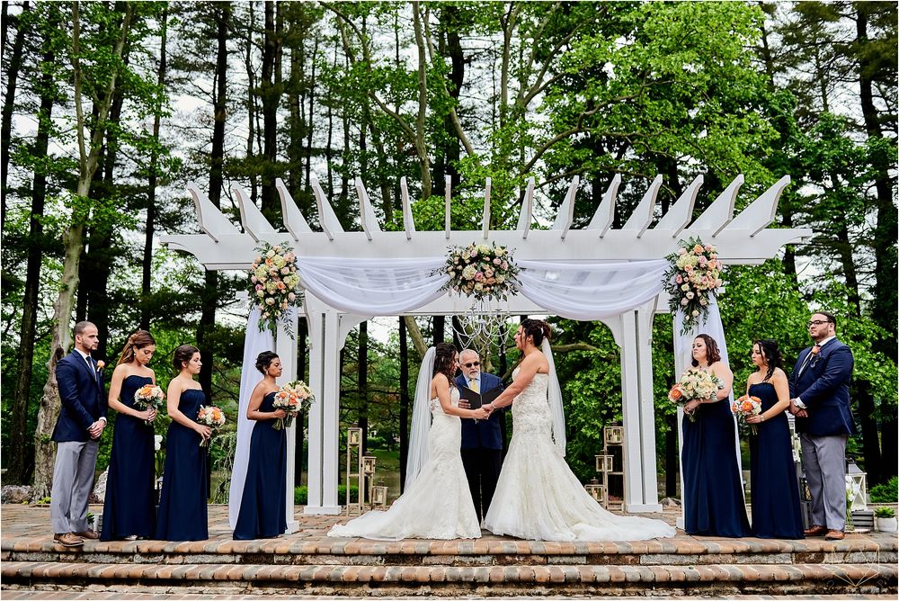 Wedding ceremony under a white pergola with bridal party in navy blue dresses standing in formation.