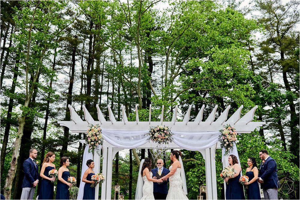 Outdoor wedding ceremony with elegant white pergola and wedding party in navy attire against forest backdrop.