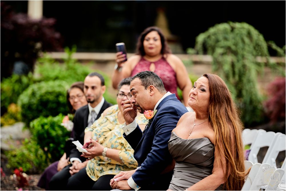 Wedding guests watching ceremony intently while seated in garden setting.