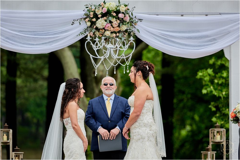 Two brides stand under an elegant floral arch during their outdoor wedding ceremony.