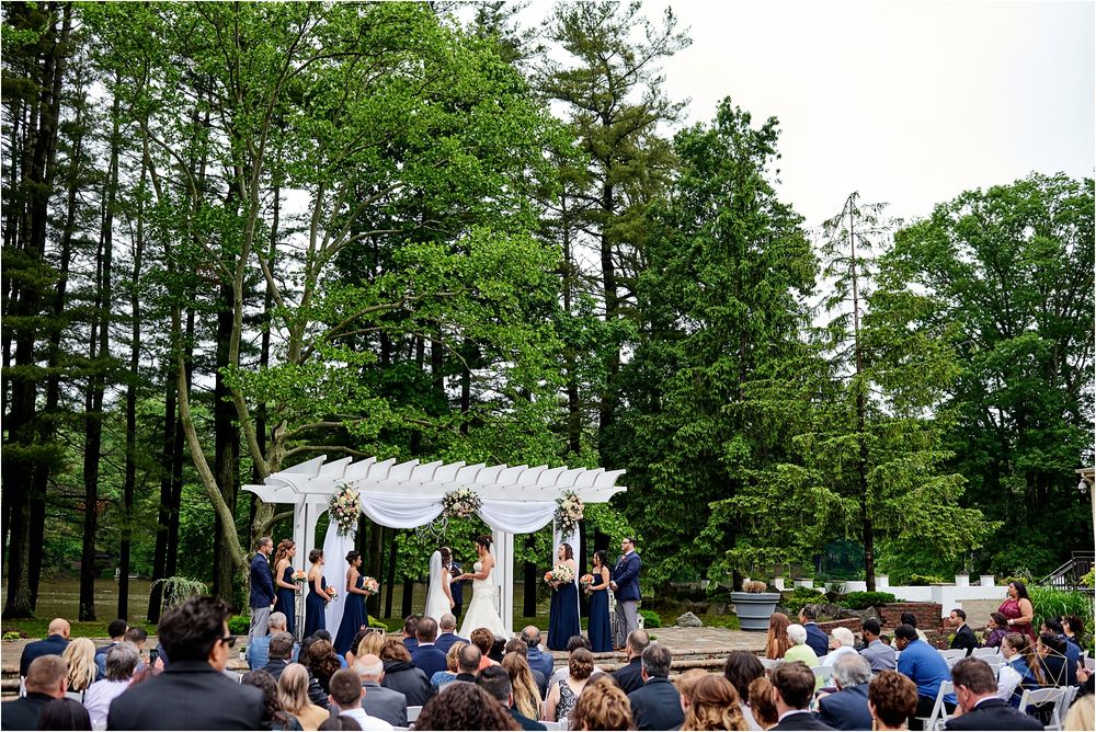 Outdoor wedding ceremony setup with white pergola and rows of chairs surrounded by tall green trees.