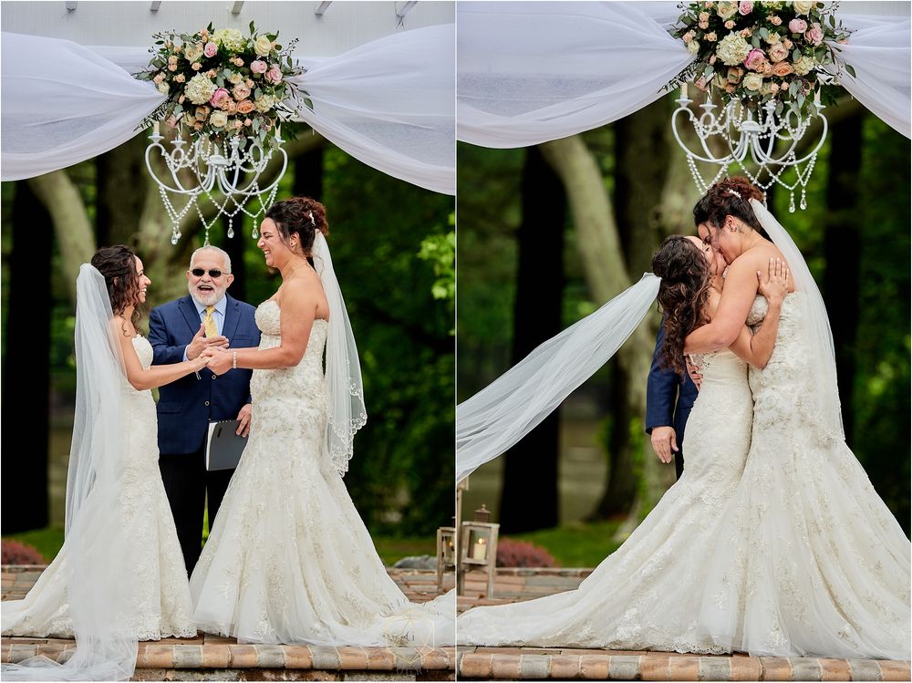 Two brides in lace wedding dresses share their first kiss during an outdoor ceremony under a crystal chandelier.