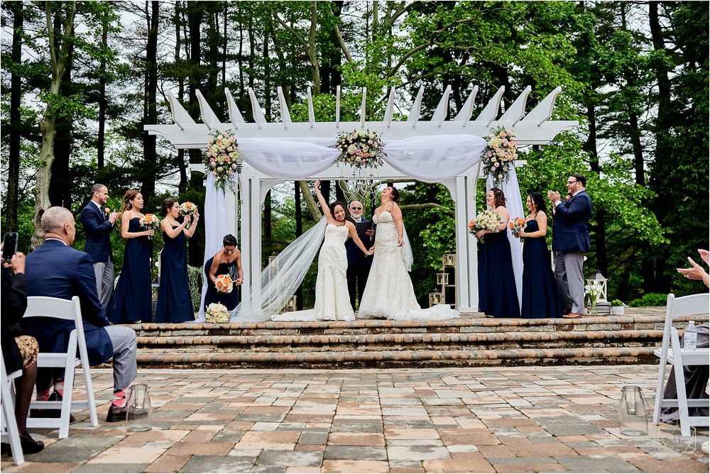 Wedding ceremony under white pergola with navy-clad wedding party and guests seated on brick patio.