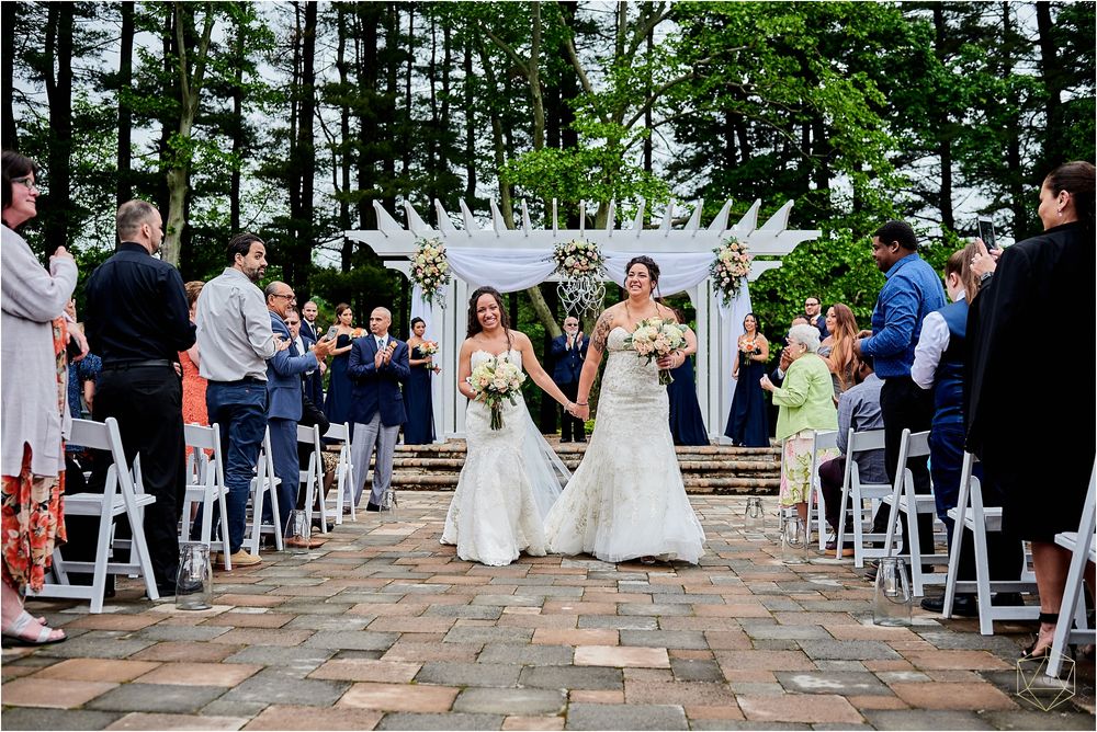 Wedding ceremony on brick patio with white pergola and guests seated in white chairs.