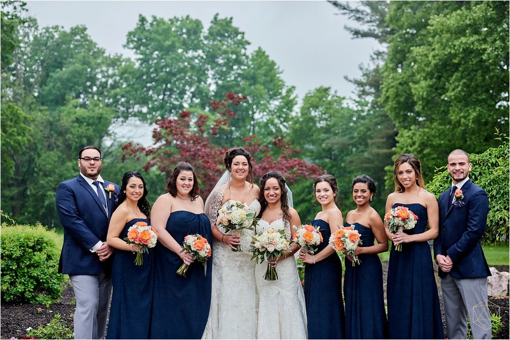 Wedding party in navy blue attire and white dresses poses outdoors in garden setting.