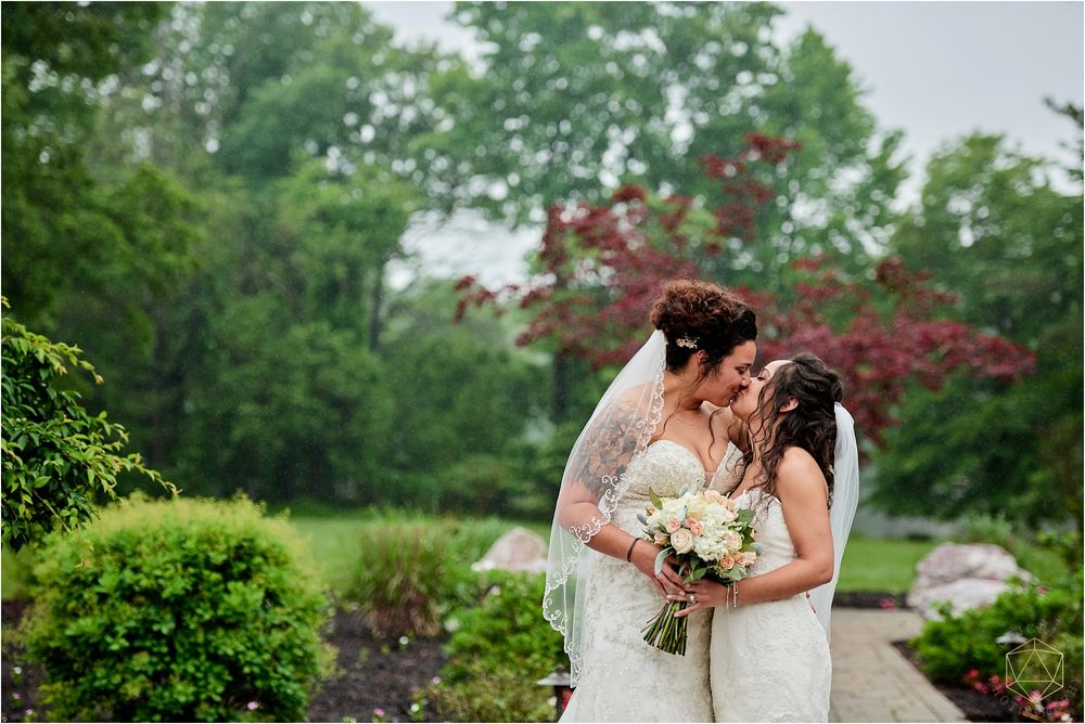 Two brides share intimate moment under blooming Japanese maple tree in garden setting.