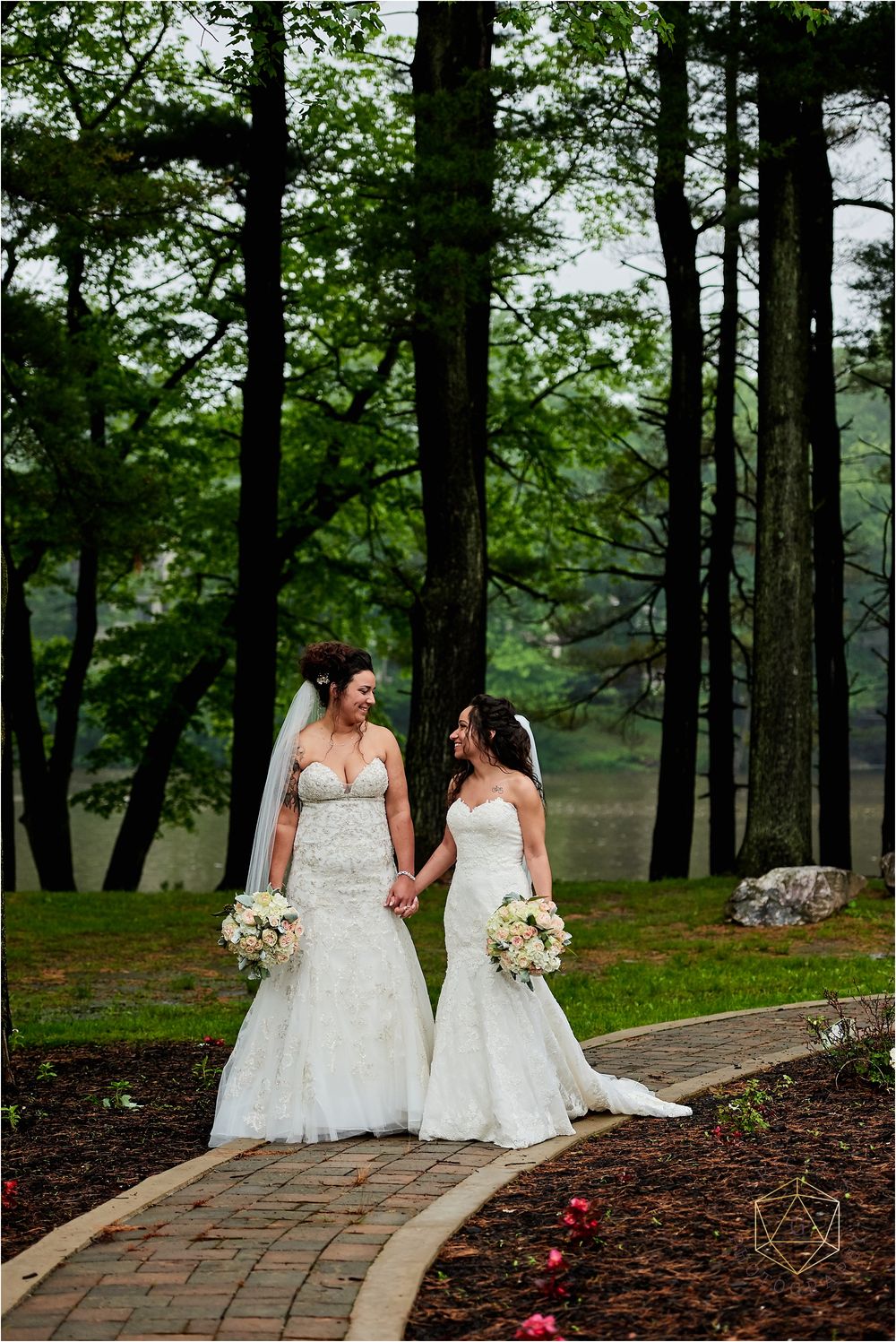 Two brides in lace gowns hold hands while walking stone path through pine forest.