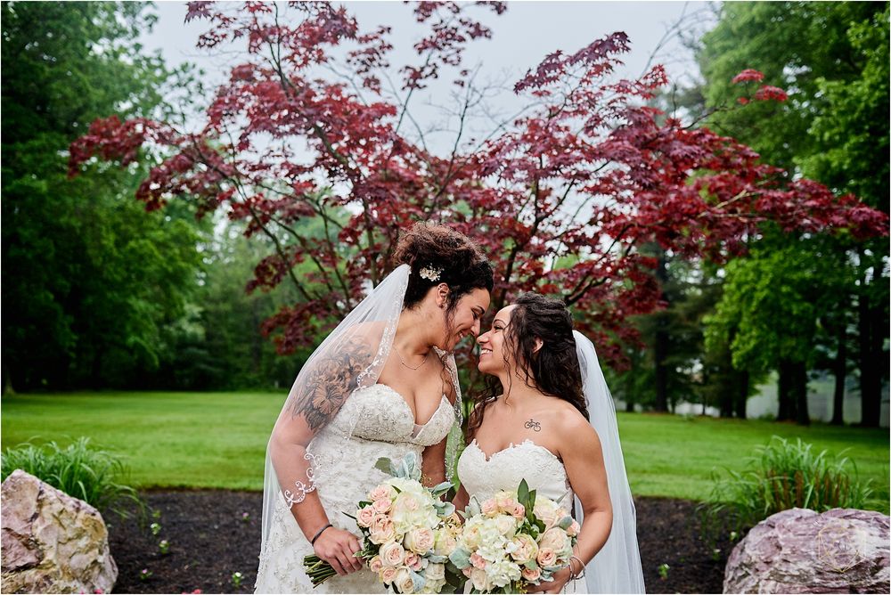 Two brides with white bouquets pose under red Japanese maple tree in spring garden.
