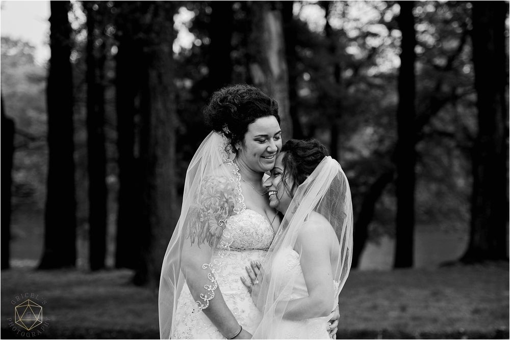 Two brides share an intimate moment in a forest setting captured in black and white.