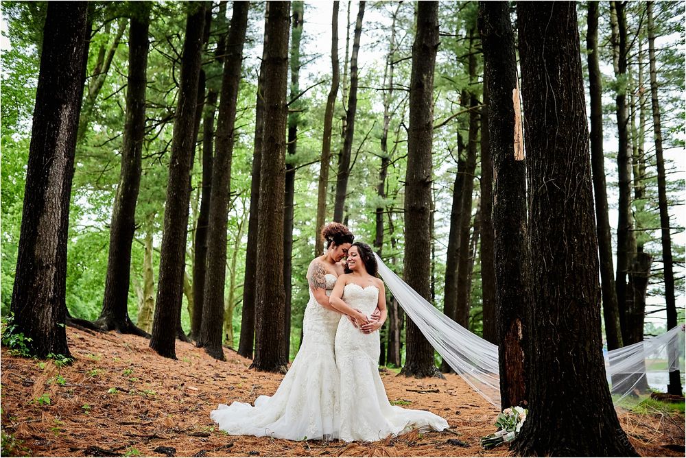 Two brides in white gowns embrace among tall trees in a romantic forest setting.