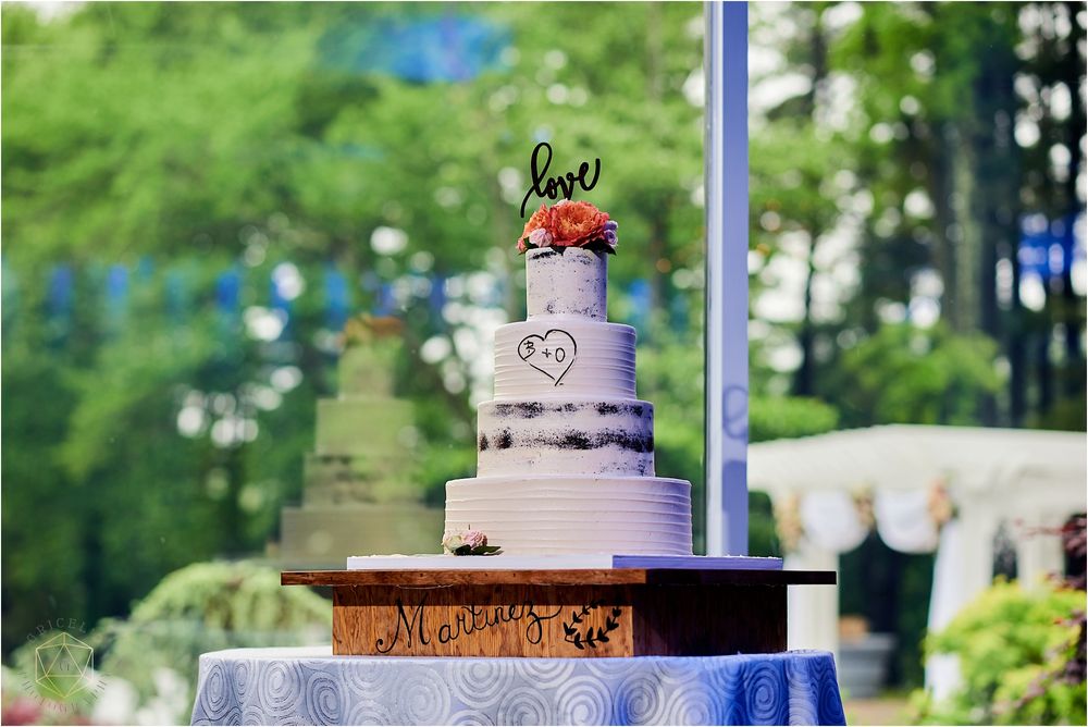 White wedding cake with red floral topper displayed outdoors against blurred greenery.