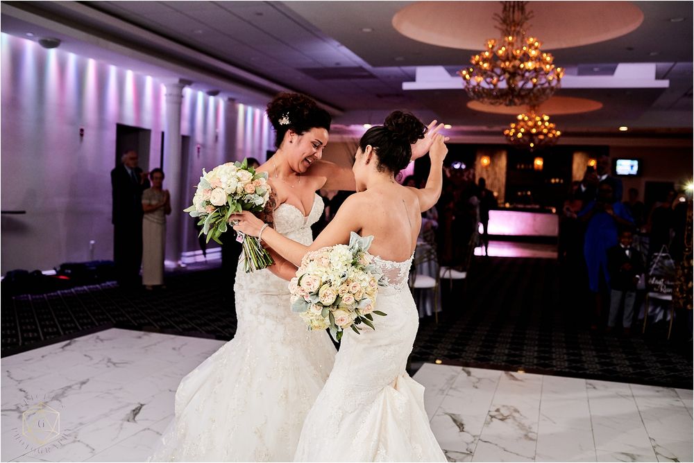 Two brides dancing together in an elegant ballroom with purple accent lighting.