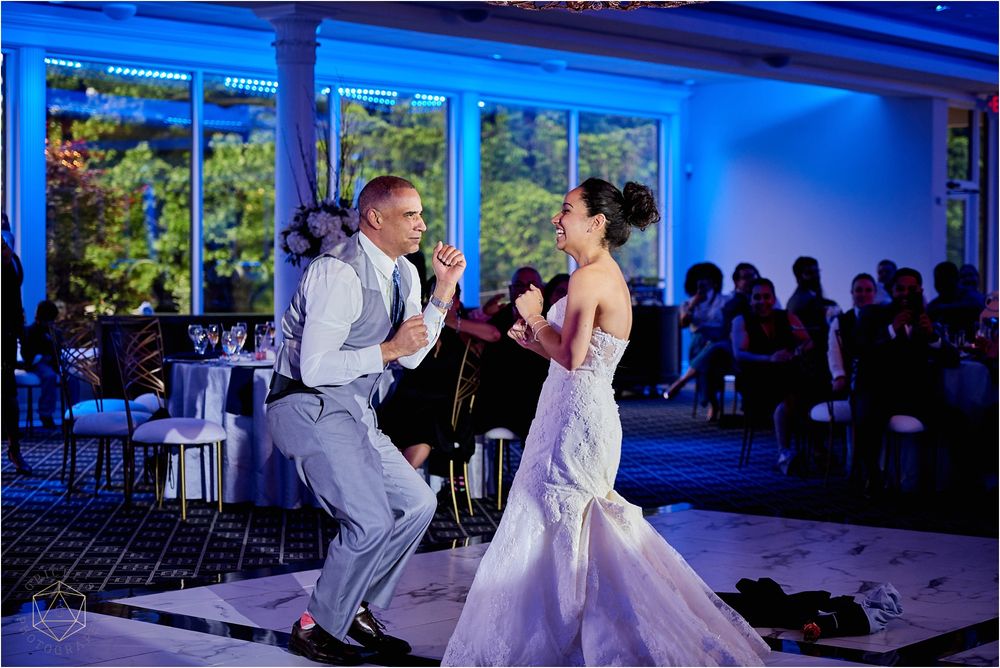 A couple performs an energetic dance routine on their wedding day in a blue-lit reception hall.