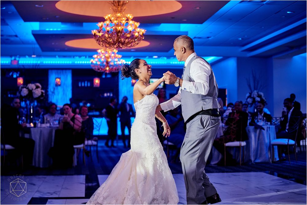 A couple dances joyfully under a crystal chandelier in a blue-lit ballroom at their wedding reception.