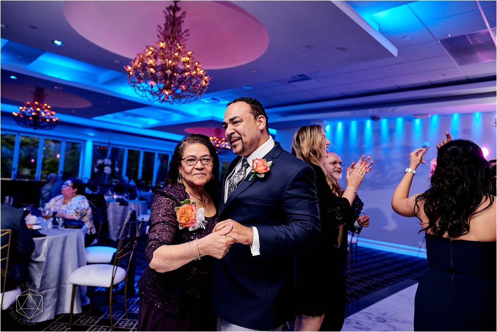 Wedding guests dance together under blue lighting and crystal chandeliers at an elegant reception venue.