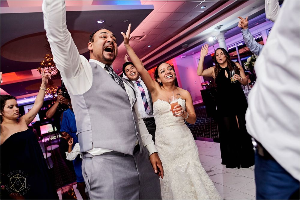 Wedding reception dance party with guests celebrating enthusiastically under pink and purple lighting.